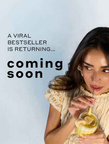 Model with brown hair and french manicure wearing yellow shirt and yellow earrings holding straw of glass with yellow smoothie sliced bananas to mouth, on blue background with black text that reads "A Viral Bestseller Is Returning... Coming Soon"