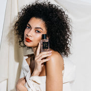 Modèle avec des cheveux noirs bouclés, un vernis à ongles nude, un rouge à lèvres rouge, enveloppée dans un drap blanc contre un fond en tissu blanc, tenant le parfum Myth 50 ml sur l'épaule.