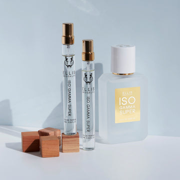 Iso Gamma Super 10ml Eau De Parfum Travel Spray on wooden block between three other wooden blocks on table and Iso Gamma Super 10ml Eau De Parfum Travel Spray on table, next to Iso Gamma Super 50ml Eau De Parfum on table, on light grey background.