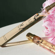Two Raven 10ml Eau De Parfum Travel Sprays next to a pink peony flower, on white table, against grey background