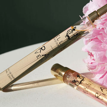 Two Raven 10ml Eau De Parfum Travel Sprays next to a pink peony flower, on white table, against grey background