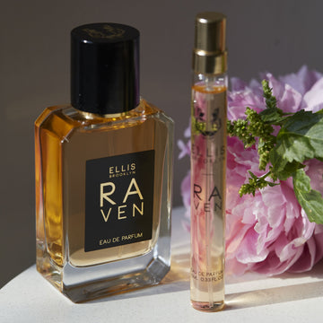 Raven 50ml Eau De Parfum on white table next to Raven 10ml Eau De Parfum Travel spray, pink flower, and green leaves, on brown background.