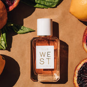 West 50ml Eau De Parfum on textured light brown background surrounded by blood oranges, basil, and lemons.
