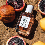 West 50ml Eau De Parfum on dirt next to blood oranges and lemon.